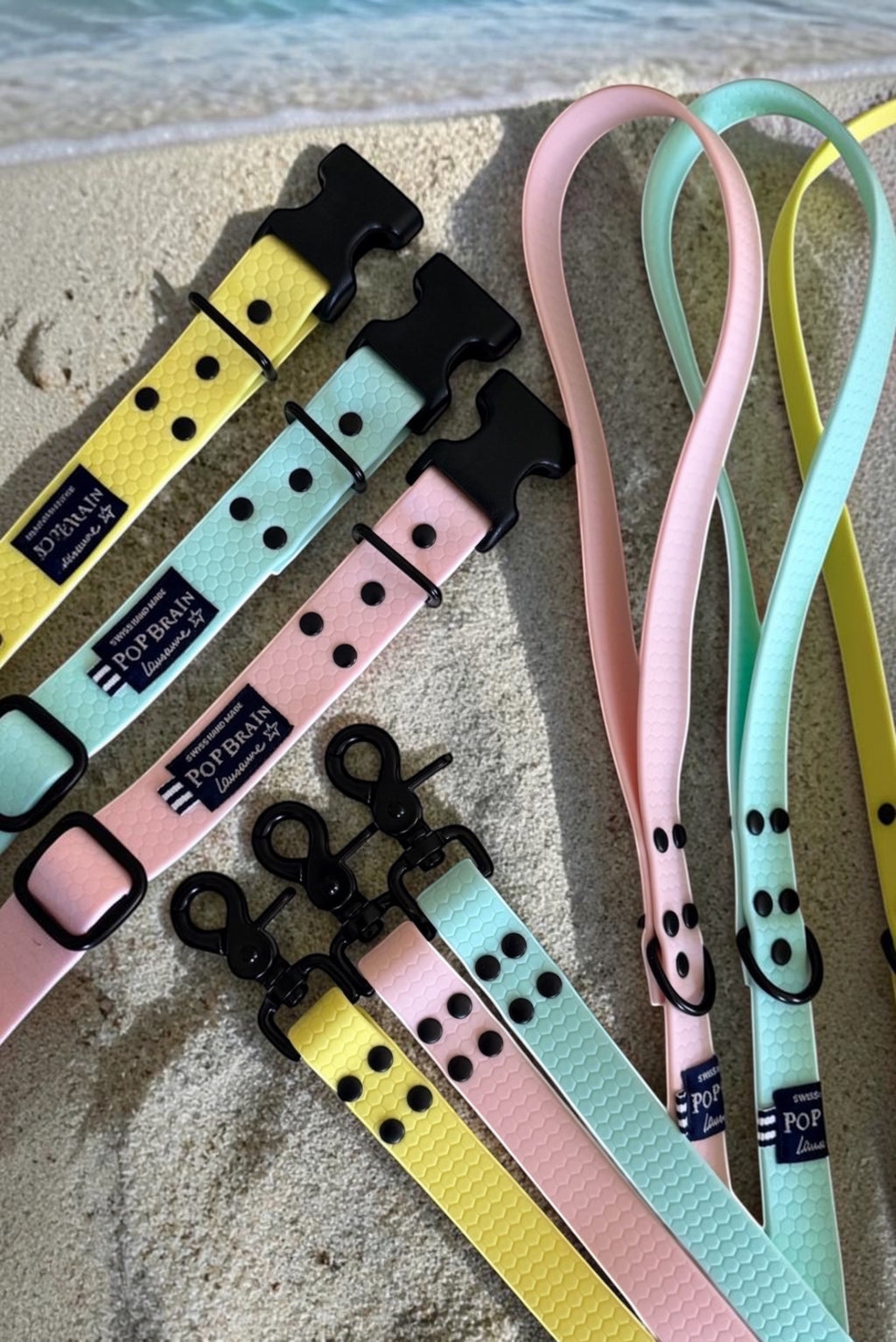 Popbrain waterproof PVC dog leashes and collars in pastel yellow, pink, and mint with matte black hardware on sand.